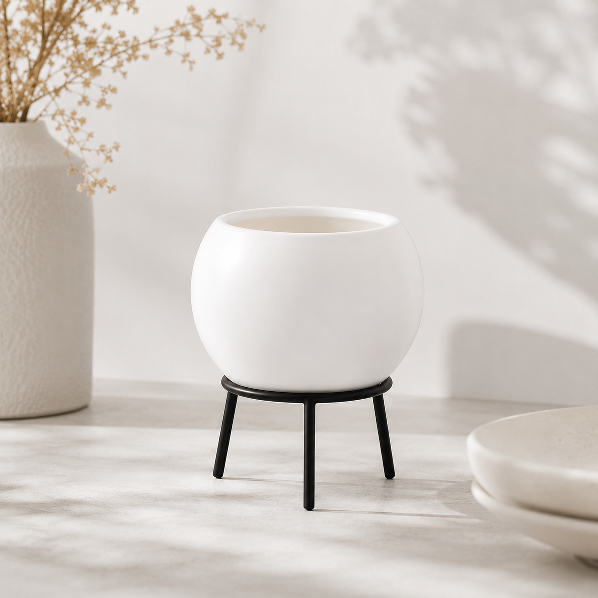A matte white round planter pot resting on a black metal stand with three legs, set on a light stone surface with soft shadows and a decorative vase in the background.