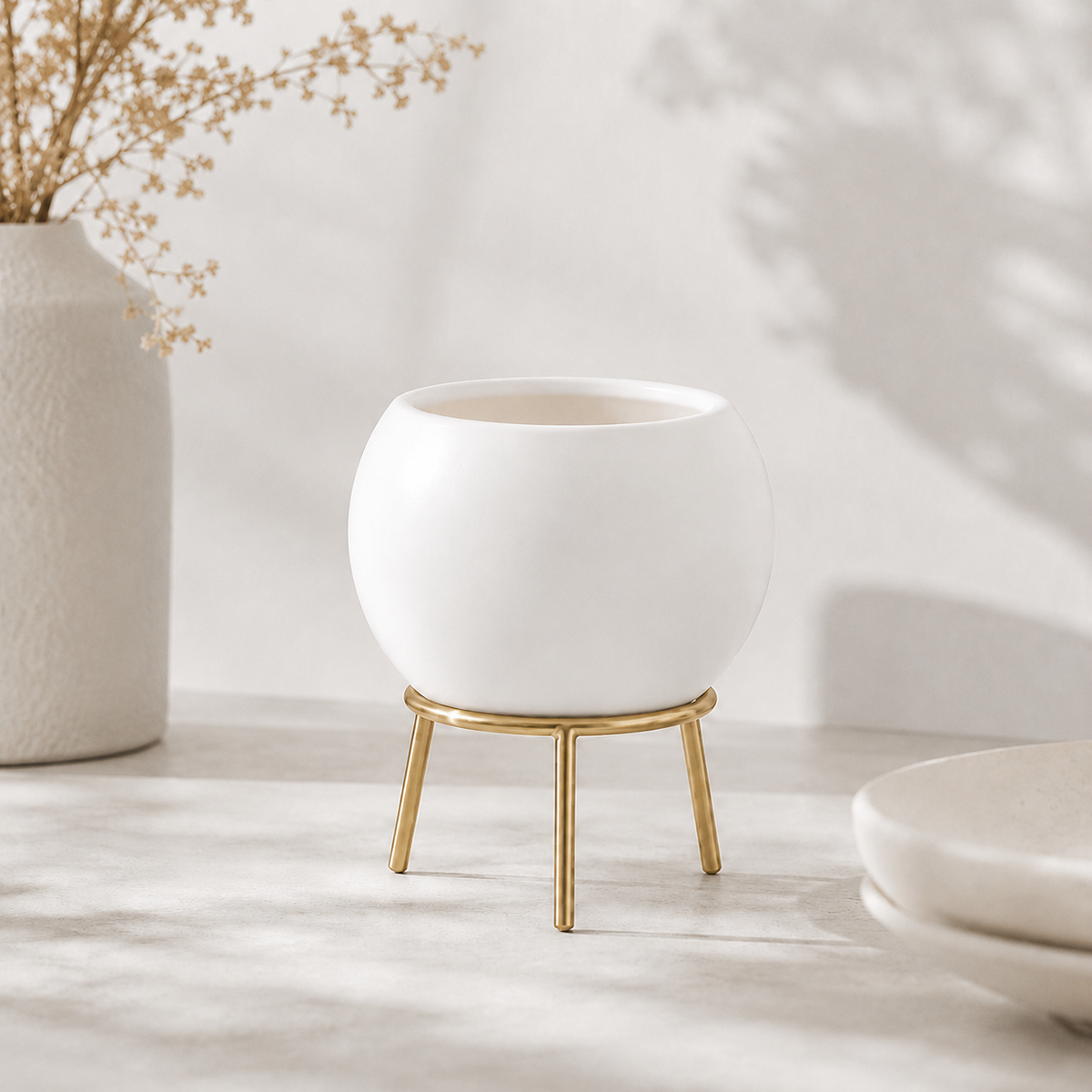 A white ceramic planter pot with a small gold metal stand, styled on a light marble surface with soft shadows and a textured vase in the background.