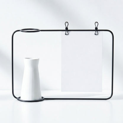 A minimalist black metal desk organizer frame with two hanging clips holding blank white paper, styled with a white ceramic vase on a clean white surface against a white background.