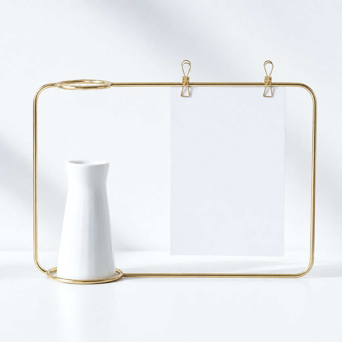 A gold metal wire frame desk organizer with two hanging clips holding a blank white sheet of paper, styled with a small white ceramic vase on a white surface against a white background.