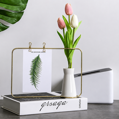 A gold metal clip frame stand holding a printed card with a green leaf illustration, styled on top of a white book with black script. A small white vase with pink and white tulips sits in the center, with a white box and a large green leaf in the background against a light gray wall.