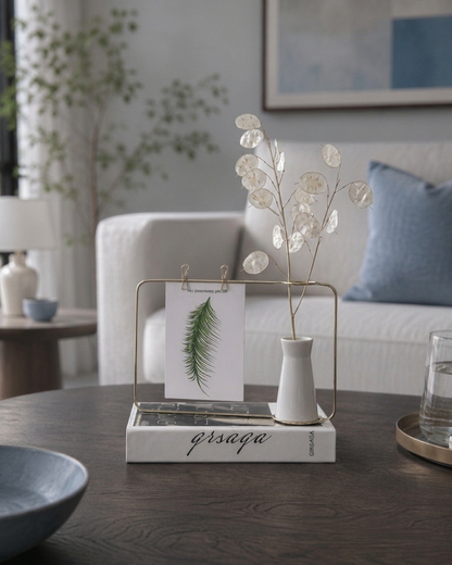A styled home decor scene showing a gold metal frame organizer holding a small white vase with round leafy stems and a card, placed on a white book on a dark wooden table in a living room setting.
