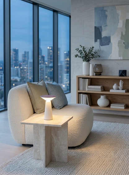 A modern living room corner featuring a rounded cream boucle armchair with two pillows, a matching light wood side table with a glowing sculptural table lamp, and a light rug. Large floor-to-ceiling windows show a city skyline at dusk, and a wooden shelf with decorative vases and books sits against the wall under abstract wall art.