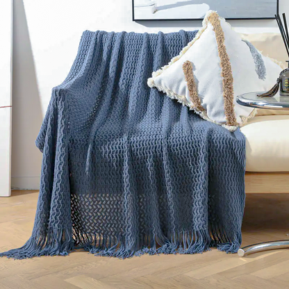 A textured blue throw blanket with fringe is draped over a chair in a bright living room setting, styled with a white and beige decorative pillow.
