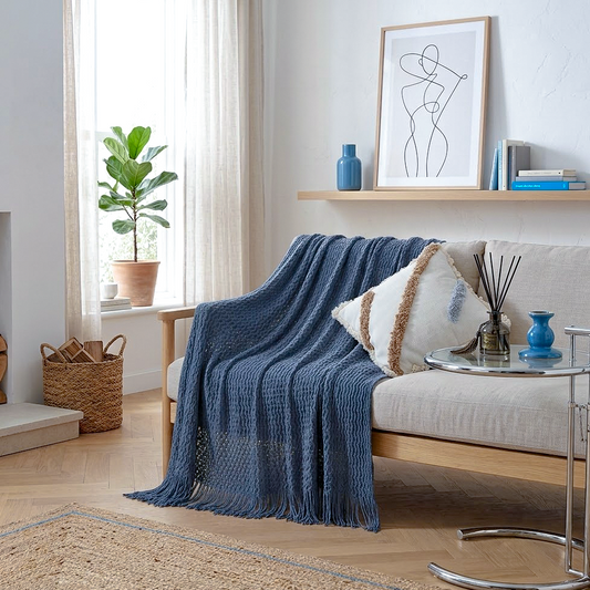 A textured navy blue knit throw blanket with fringe is draped over a light beige sofa in a bright, modern living room. The scene includes a wooden shelf with a blue vase and framed art, a potted plant, and a small side table.