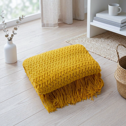 A folded mustard yellow textured throw blanket with fringe trim sits on a light wood floor in a bright, modern room setting.
