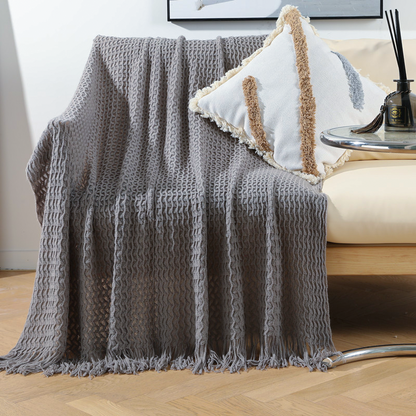 A textured gray knit throw blanket with fringe is draped over a cream-colored armchair in a bright, modern living room setting.