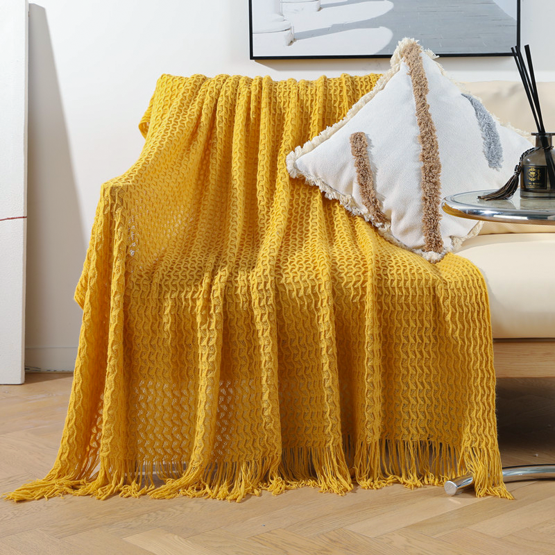 A mustard yellow textured knit throw blanket with fringe is draped over a light beige armchair in a bright living room setting, styled with a white decorative pillow.