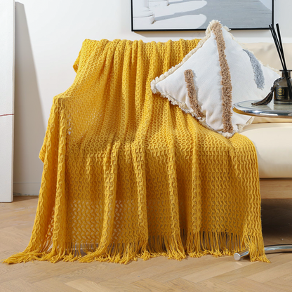 A mustard yellow textured knit throw blanket with fringe is draped over a light beige armchair in a bright living room setting, styled with a white decorative pillow.