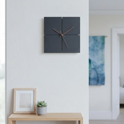 A modern four-panel square wall clock in dark charcoal gray with thin brown hands, mounted on a white wall above a light wood console table with a small potted succulent and a picture frame.