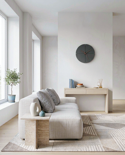 A modern living room featuring a light beige textured sofa with gray throw pillows, a matching stone side table, a geometric patterned rug, and a minimalist light wood console table against a white wall with a round dark gray wall clock.