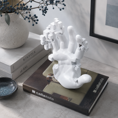 A white sculptural hand-shaped decor object with geometric tree-like branches, displayed on top of a book in a styled interior setting with books, a vase, and framed wall art.