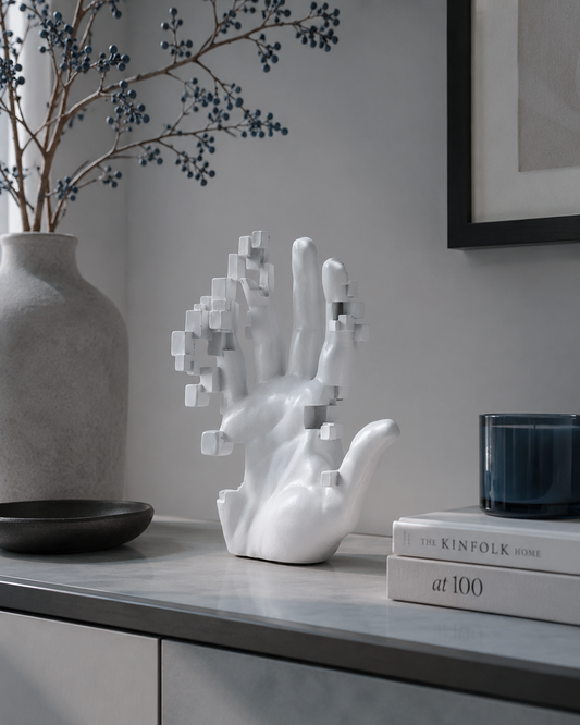 A white sculptural hand sculpture with pixelated cube details on the fingers, displayed on a gray console table in a modern interior setting next to a gray vase with dried blue berries, a black bowl, books, and a dark blue candle jar.