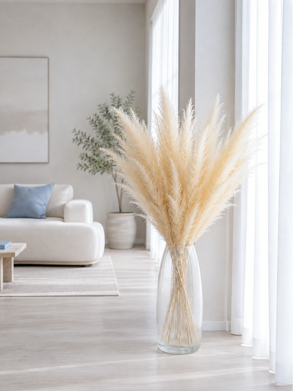 A large vase filled with tall beige dried reed grass stands on a light wood floor in a bright, modern living room with a white sofa and large windows.