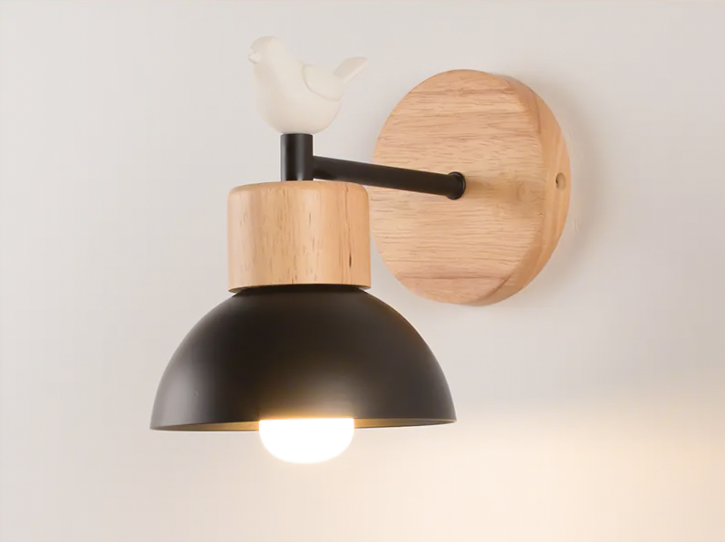 A modern wall-mounted light fixture with a matte black dome shade, natural wood accents, and a white bird sculpture on top, shown lit against a plain light background.