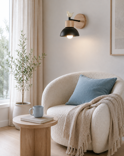 A modern wall-mounted sconce with a black dome shade and a light wood backplate, shown lit in a Scandinavian reading nook next to a cream boucle armchair with a blue pillow and beige throw blanket.