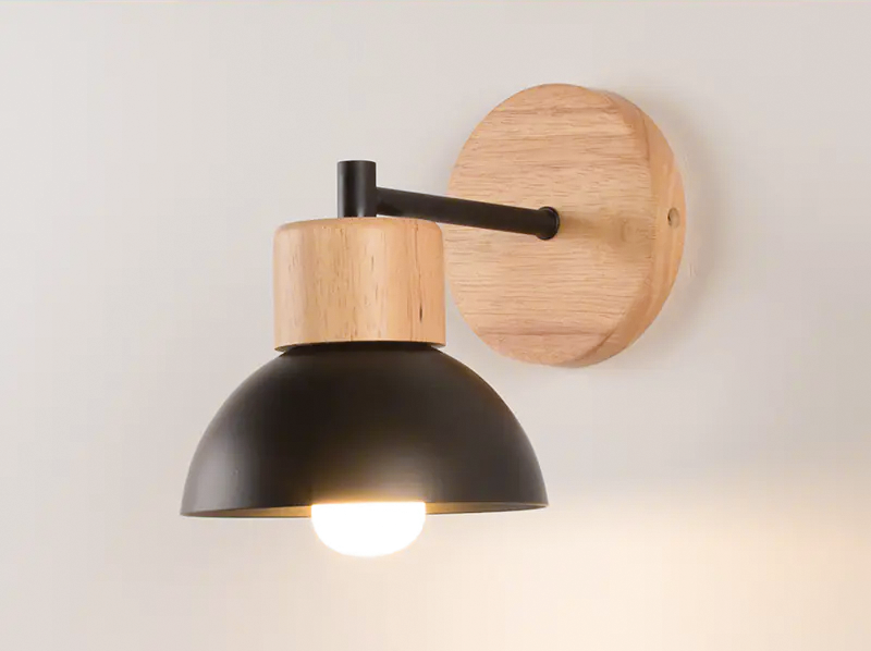 A modern wall-mounted light fixture with a black dome-shaped shade, a light natural wood neck and backplate, and a black metal arm, shown lit against a plain off-white wall.