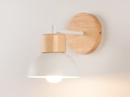 A modern wall-mounted light fixture with a white dome-shaped shade, a light natural wood neck, and a round natural wood backplate, shown lit against a plain off-white wall.