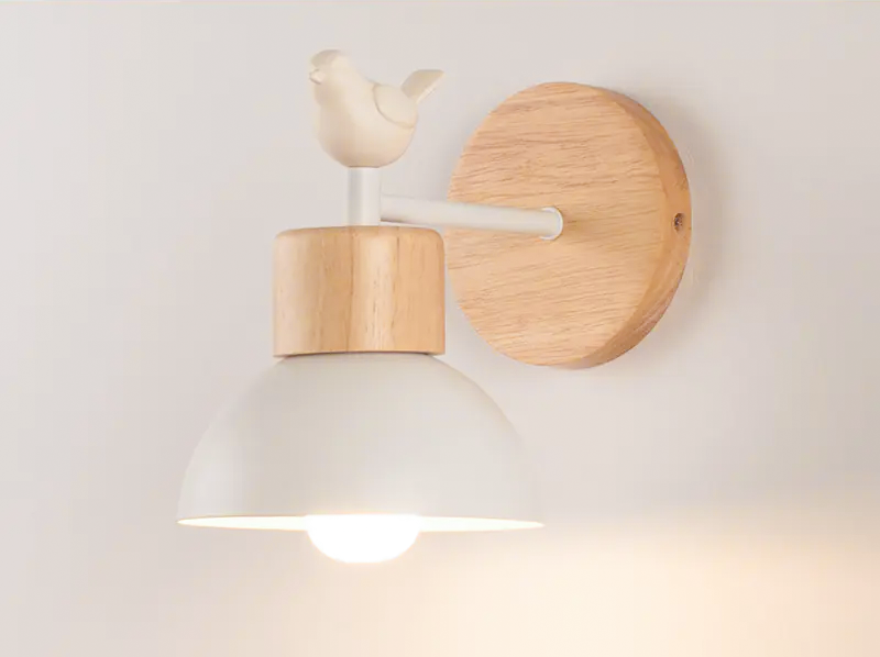 A wall-mounted light fixture with a white dome-shaped shade, a light natural wood neck and circular wall plate, and a small bird sculpture on top, shown lit against a plain light wall.