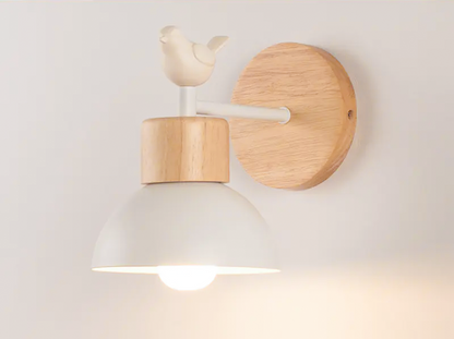 A wall-mounted light fixture with a white dome-shaped shade, a light natural wood neck and circular wall plate, and a small bird sculpture on top, shown lit against a plain light wall.