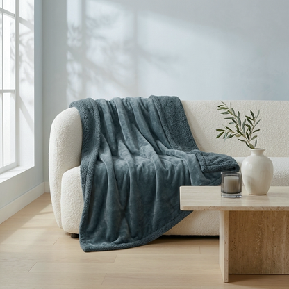 A plush, dusty blue throw blanket is draped over a cream boucle sofa in a bright, modern living room setting with a wooden coffee table and vase.