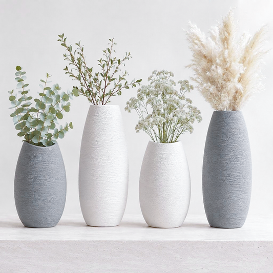 Four textured ceramic vases in gray and white, arranged on a light surface against a white background, with green and beige dried botanicals placed inside.