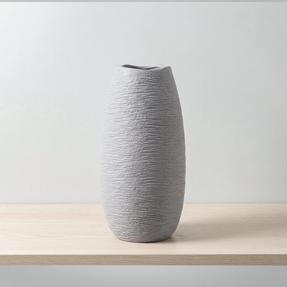 A tall, textured tall gray ceramic vase with a small opening, standing on a light wood surface against a neutral light gray background.
