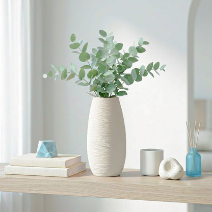 A tall textured off-white ceramic vase filled with eucalyptus stems, styled on a light wood shelf with books, a small blue geometric object, a gray cup, a white decorative stone, and a blue reed diffuser in a bright interior setting.