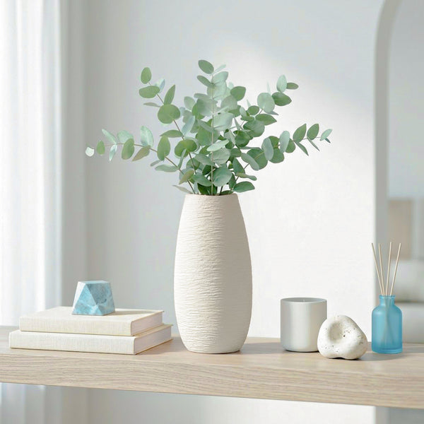 A tall textured off-white ceramic vase filled with eucalyptus stems, styled on a light wood shelf with books, a small blue geometric object, a gray cup, a white decorative stone, and a blue reed diffuser in a bright interior setting.