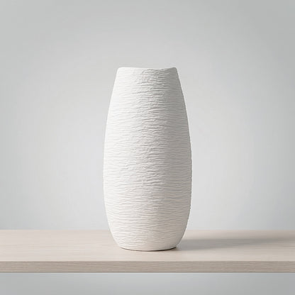 A tall, textured white ceramic vase with a flared rim, displayed on a light wood shelf against a neutral gray background.