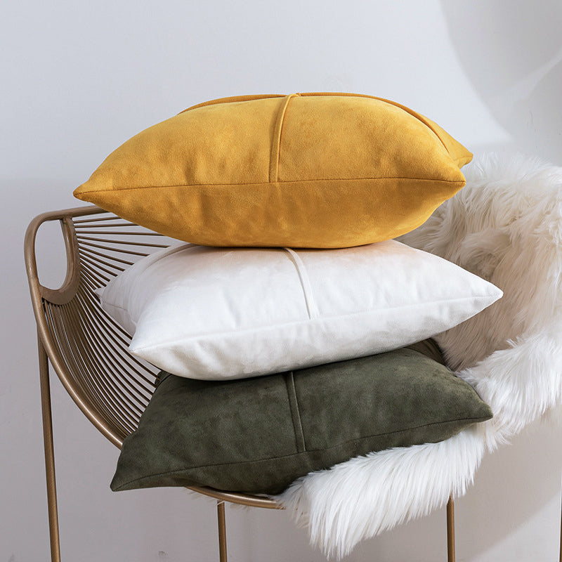 Three decorative throw pillows in mustard yellow, white, and dark olive green are stacked on a gold wire chair with a white faux-fur throw against a light gray wall.