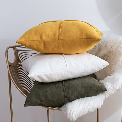 Three decorative throw pillows in mustard yellow, white, and dark olive green are stacked on a gold wire chair with a white faux-fur throw against a light gray wall.