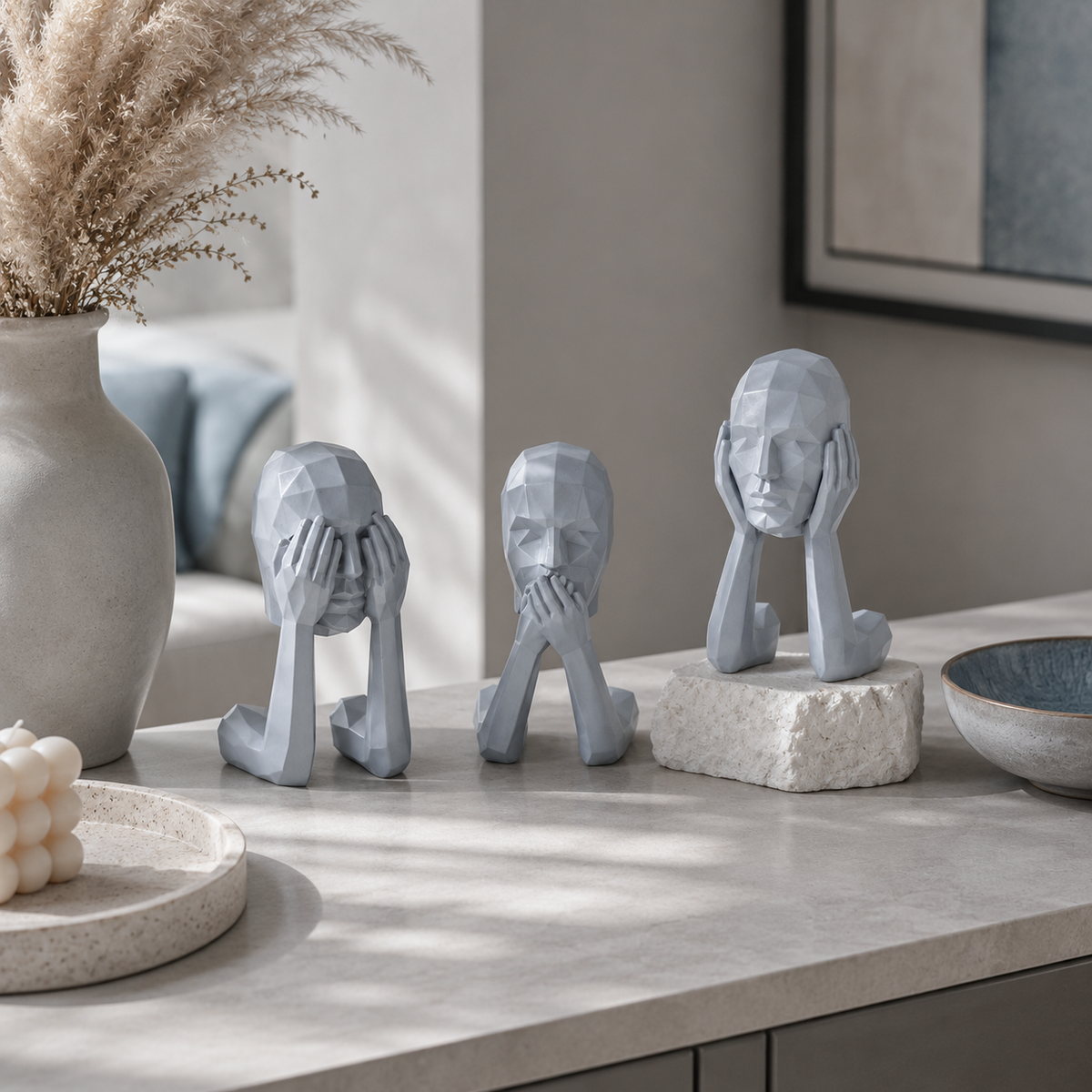 Three gray geometric figurines of stylized human heads and hands arranged on a light stone countertop in a modern interior setting.