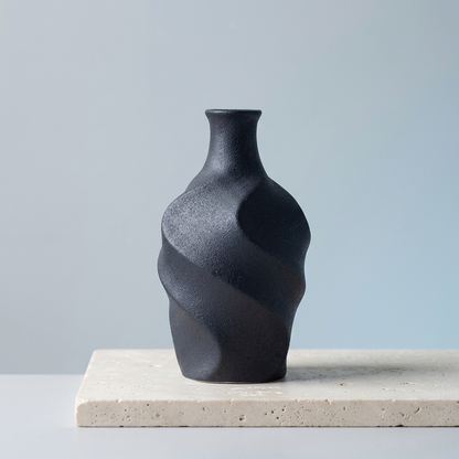 A small matte black ceramic vase with a sculptural, twisted spiral design, displayed on a light beige stone slab against a soft light gray background.