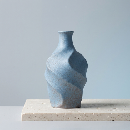A small blue ceramic vase with a textured, sculptural spiral design, displayed on a light beige stone slab against a soft light blue-gray background.