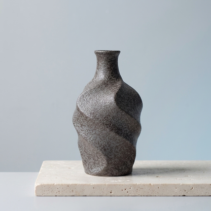 A small dark brownish gray ceramic vase with a textured, organic wavy shape, displayed on a light beige stone slab against a soft light blue-gray background.