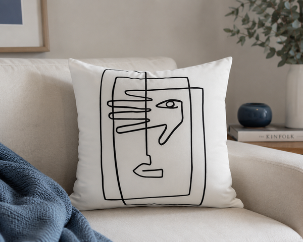 A square white throw pillow with a minimalist black line art design of a face and hand, resting on a beige sofa in a styled living room setting.