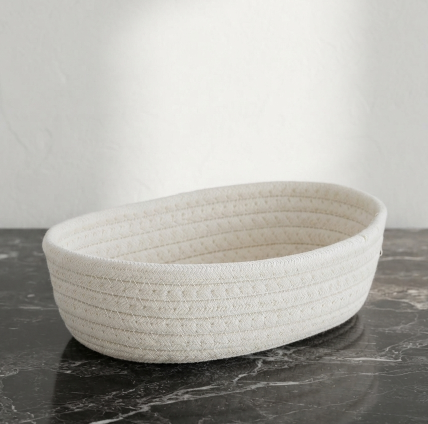 An oval woven basket made of textured off-white rope or jute material, sitting on a dark marble surface against a light gray wall.