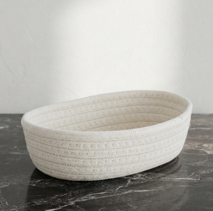 An oval woven basket made of textured off-white rope or jute material, sitting on a dark marble surface against a light gray wall.