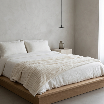 A minimalist platform bed dressed in white bedding and a cream textured throw blanket, set in a modern bedroom with textured plaster walls, a small side table, and a hanging pendant light.