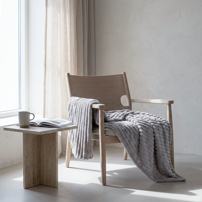 A light wood armchair in a bright, minimalist room with a chunky light gray knit throw blanket draped over it, alongside a matching side table and open book.