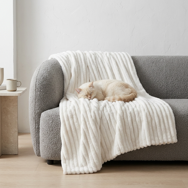 A cream-colored cat sleeps peacefully on a white ribbed fleece throw blanket draped over a gray boucle sofa in a bright, modern living room.