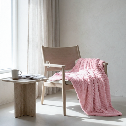 A soft pink textured throw blanket draped over a light wood armchair in a bright, minimalist room with a side table and window.