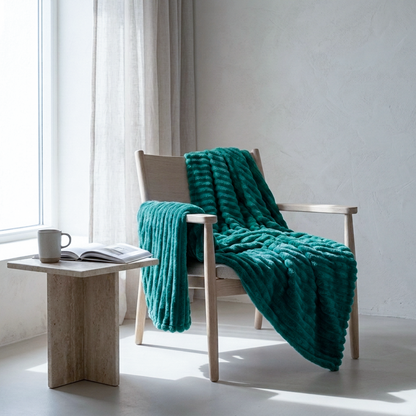 A chunky knit throw blanket in a rich viridian green color is draped over a light wood armchair in a bright, modern room. A small stone side table next to the chair holds a mug and an open book.