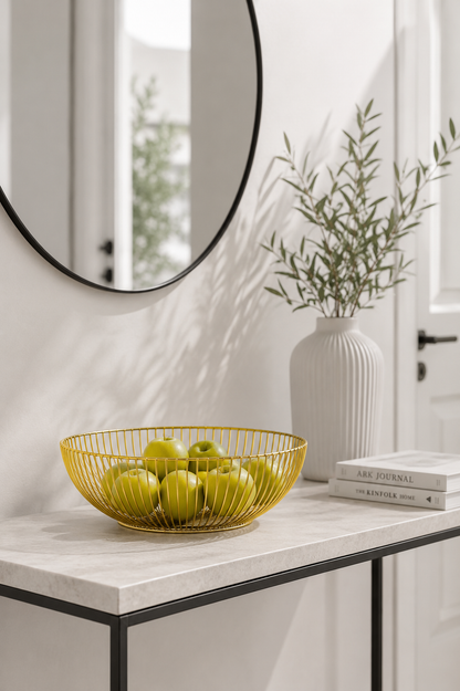 A gold wire mesh fruit bowl filled with green apples sits on a white marble console table in a bright, modern interior. A white ribbed ceramic vase with green stems and a stack of books are placed beside it, with a large round mirror on the wall behind.