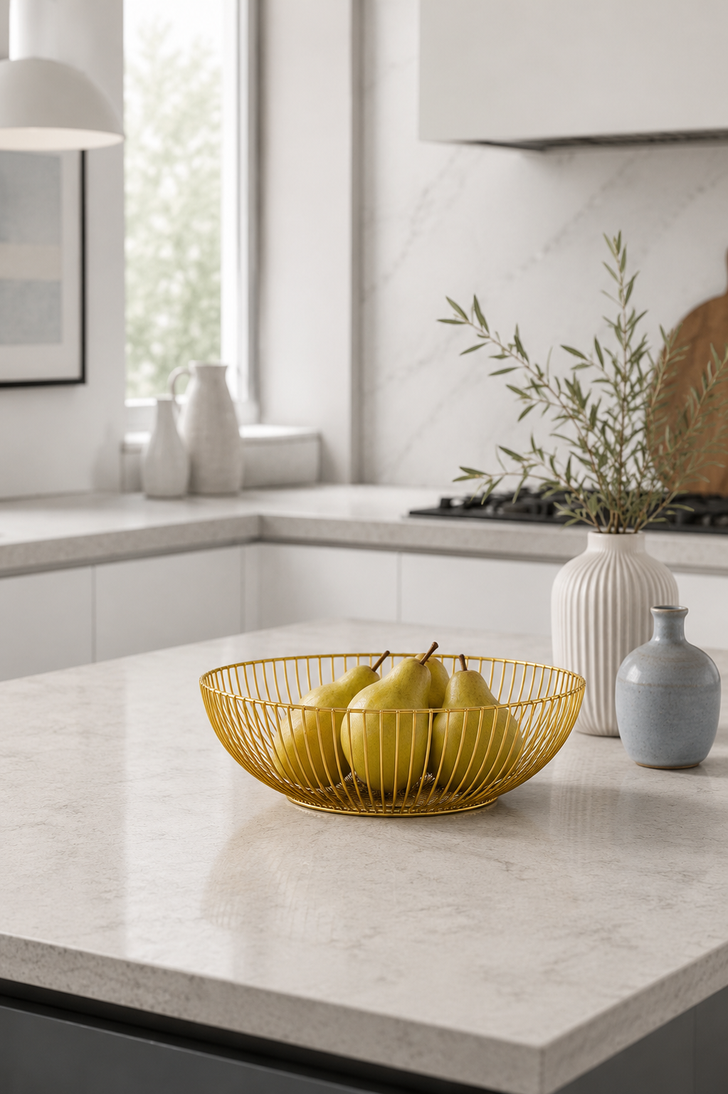A gold wire bowl filled with yellow pears sits on a light marble kitchen island, with white vases and greenery in the background.