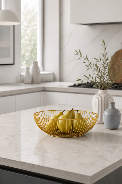 A gold wire bowl filled with yellow pears sits on a light marble kitchen island, with white vases and greenery in the background.