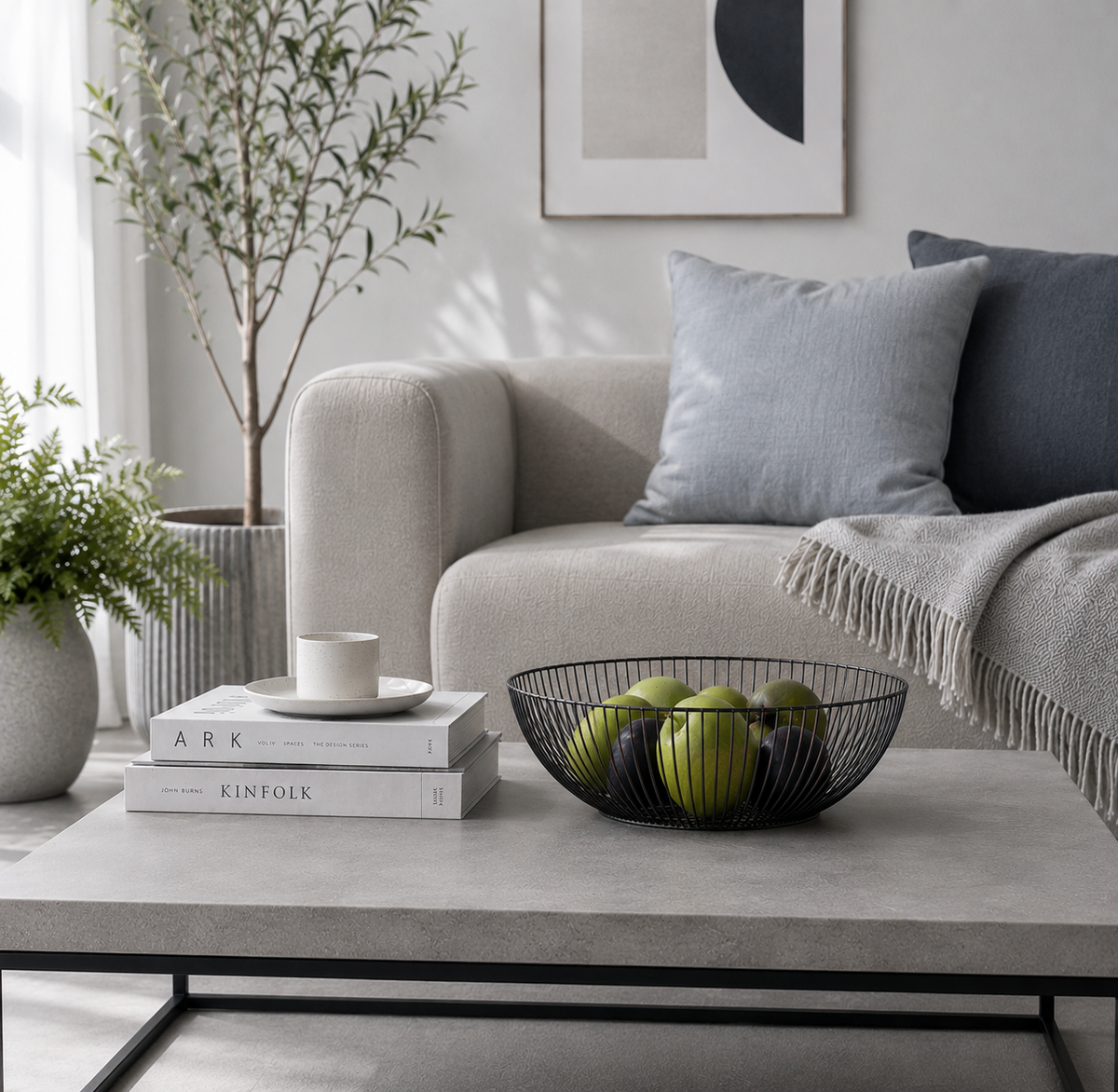 A styled living room scene featuring a light gray upholstered sofa with gray and navy throw pillows, a beige throw blanket, a gray coffee table with a  black wire fruit bowl, stacked books, and potted plants.