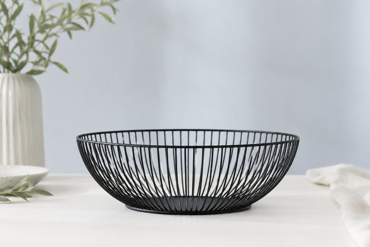 A black wire bowl with a slatted, basket-like design, placed on a white tablecloth with a neutral background and decorative props.
