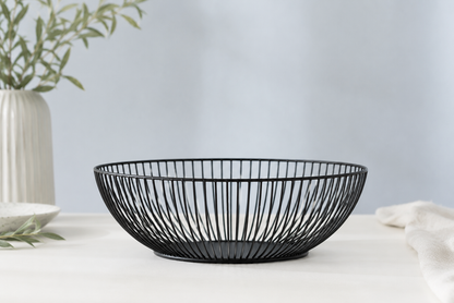 A black wire bowl with a slatted, basket-like design, placed on a white tablecloth with a neutral background and decorative props.